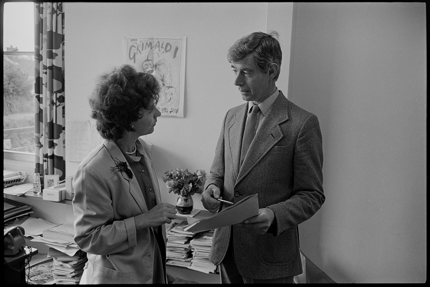 Teacher and woman secretary discussing in office. 
[A teacher and a secretary talking in an office at Chulmleigh Community College. The teacher is holding some papers and a pen.]