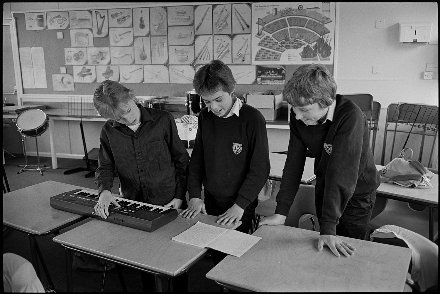 Classroom with pupils acting out short play with music. 
[Three boys reading a short play and playing a keyboard in a classroom at Chulmleigh Community College. Posters with images of musical instruments are displayed on the wall in the background.]