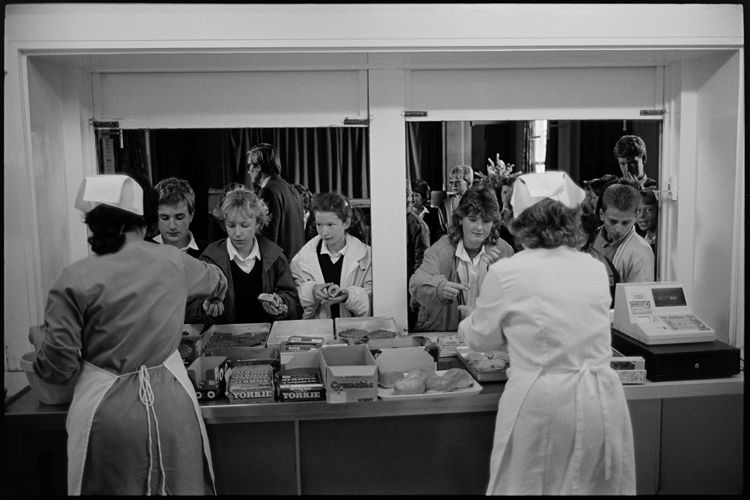 Women serving food in school canteen. 
[Two women serving food to students in the canteen at Chulmleigh Community College. Chocolate bars, pastries and a till are on the counter behind the hatch.]