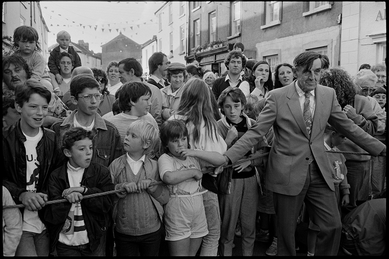 Fair Queen, woman opening fair, crowd of children at money scramble. 
[A crowd of children and adults standing behind a rope cordon waiting for the money scramble at Chulmleigh Fair, in Fore Street. A man is holding the rope.]