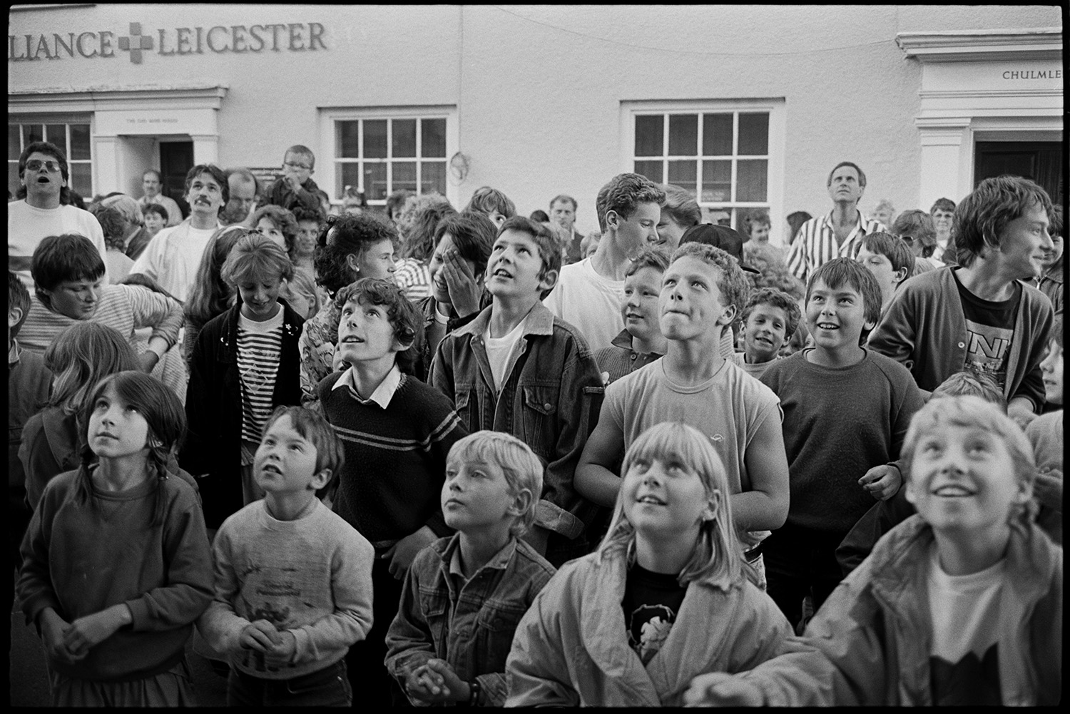 Fair, money scramble, races stalls, fancy dress. 
[Children looking up at the town hall windows waiting for money to be dropped, at the Chulmleigh Fair money scramble in Fore Street.]