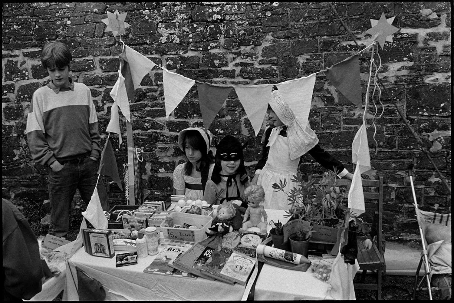 Fair, money scramble, races stalls, fancy dress. 
[Children in fancy dress running a stall selling plants, books, dolls and cassette tapes at Chulmleigh Fair. The stall is decorated with bunting.]