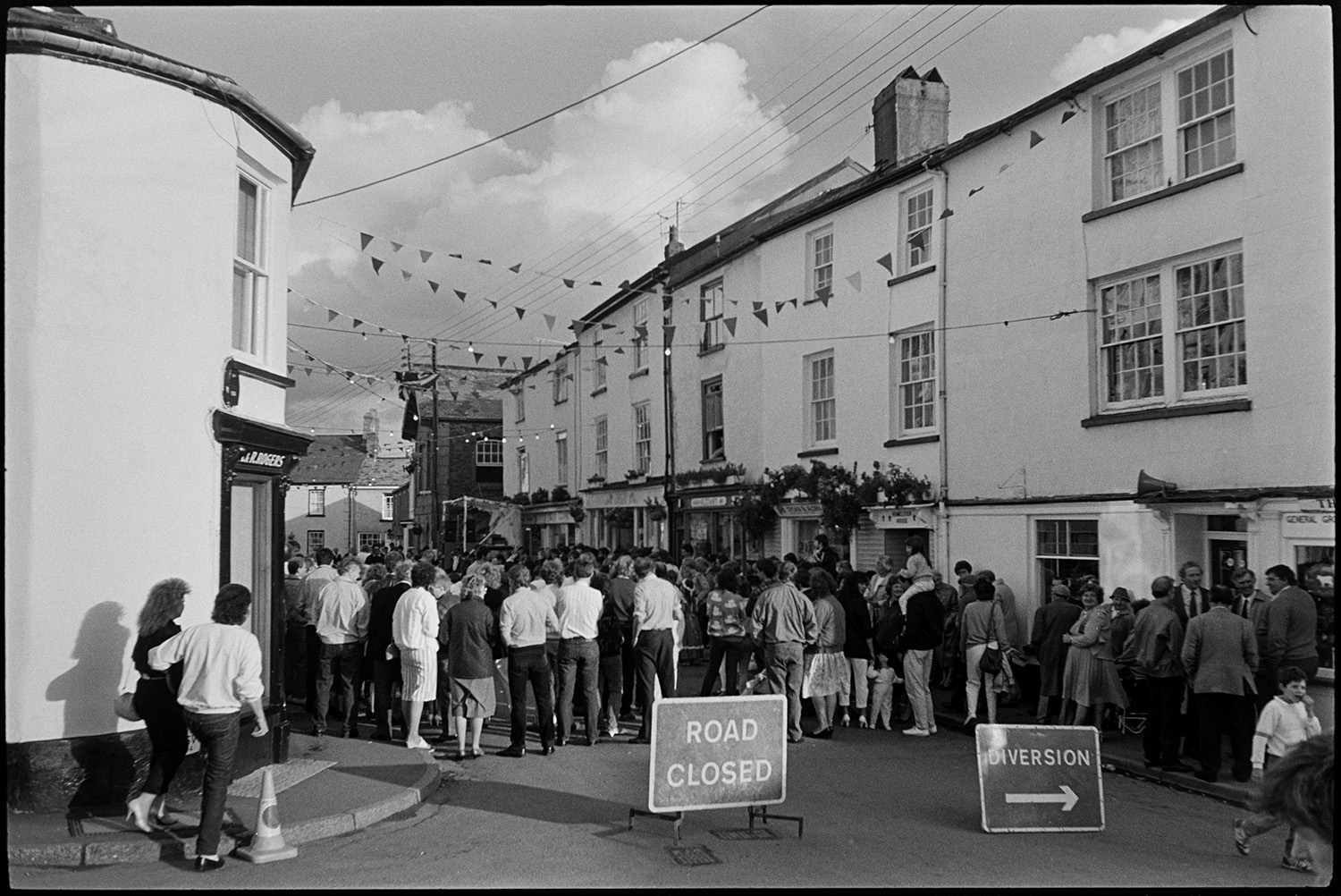 Village fair, fancy dress, crowd of onlookers, sack race. 
[Spectators gathered in a street in Chulmleigh watching events at Chulmleigh Fair. Road closed signs are visible in the foreground and the street is decorated with bunting.]