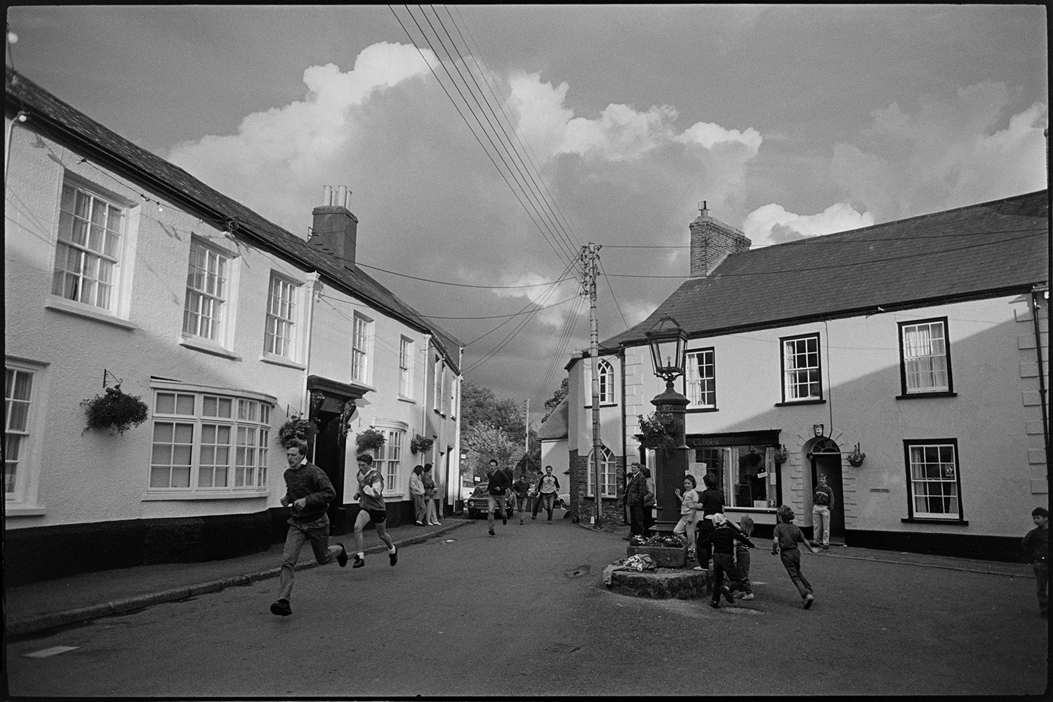 People running through a street in Chulmleigh in a race at Chulmleigh Fair. Children are watching the competitors by a lampost.