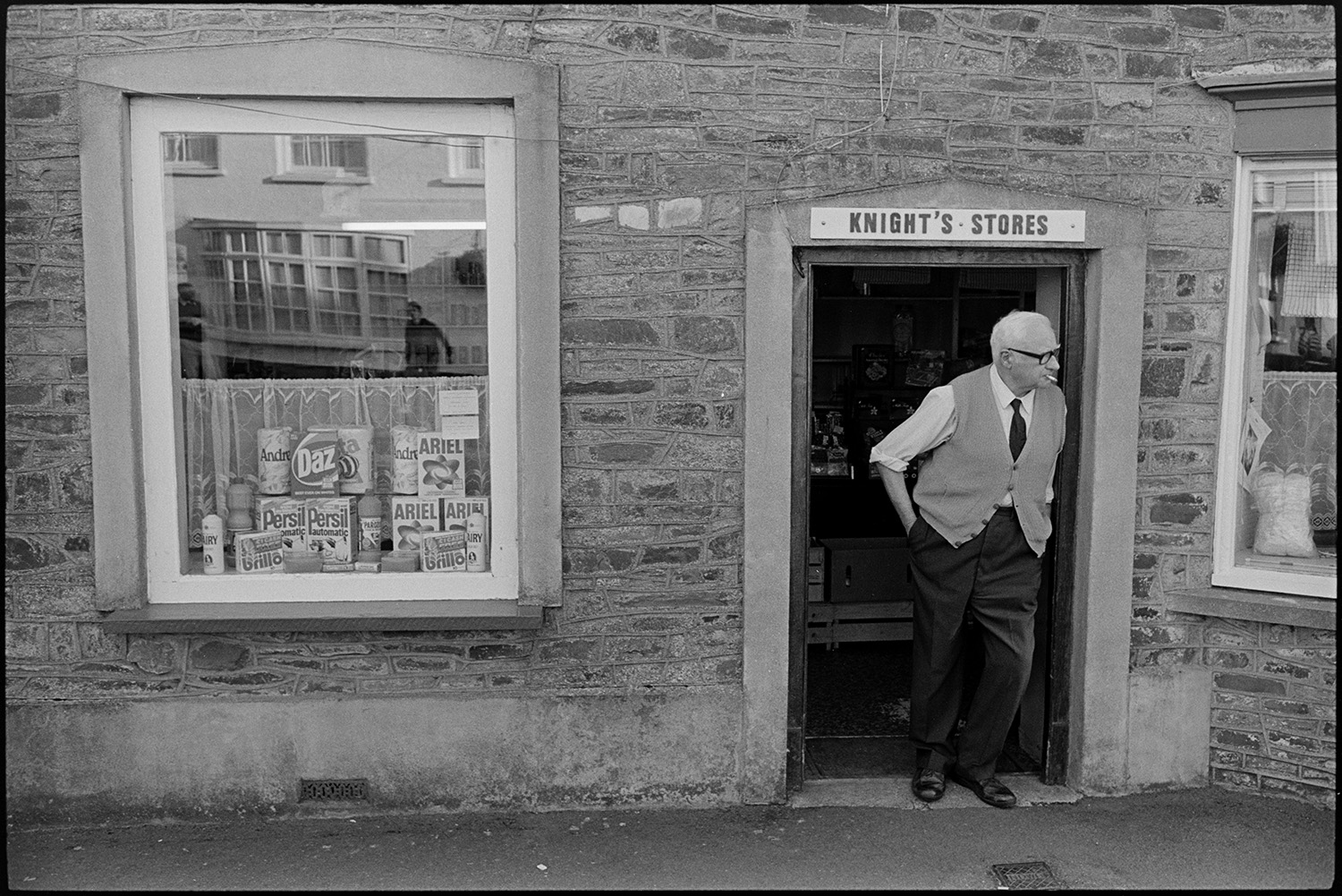Village fair, shopkeeper at shop door watching fair. 
[Bill Knight watching Chulmleigh Fair from his shop doorway of Knight's Stores. He is smoking a cigarette. Boxes of Persil, Daz and Ariel washing powder are on display in the shop window.]