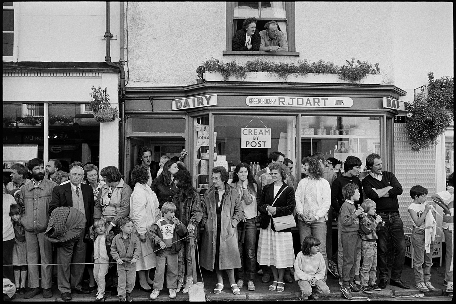 Village fair, fancy dress, crowd of onlookers, sack race. 
[Men, woman and children lined up on the pavement outside R J Dart grocers an dairy, watching events in the street at Chulmleigh Fair. Two people are also watching from the upstairs window above the shop.]