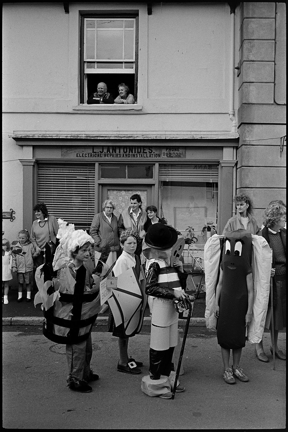Village fair, fancy dress, crowd of onlookers, sack race. 
[Children in fancy dress at Chulmleigh Fair. One child is dressed as a hot dog and another is a beacon on fire. People are watching the fancy dress parade from the side of the street, outside the shop front od L J Antunides electrical repair shop, and a first floor window.]