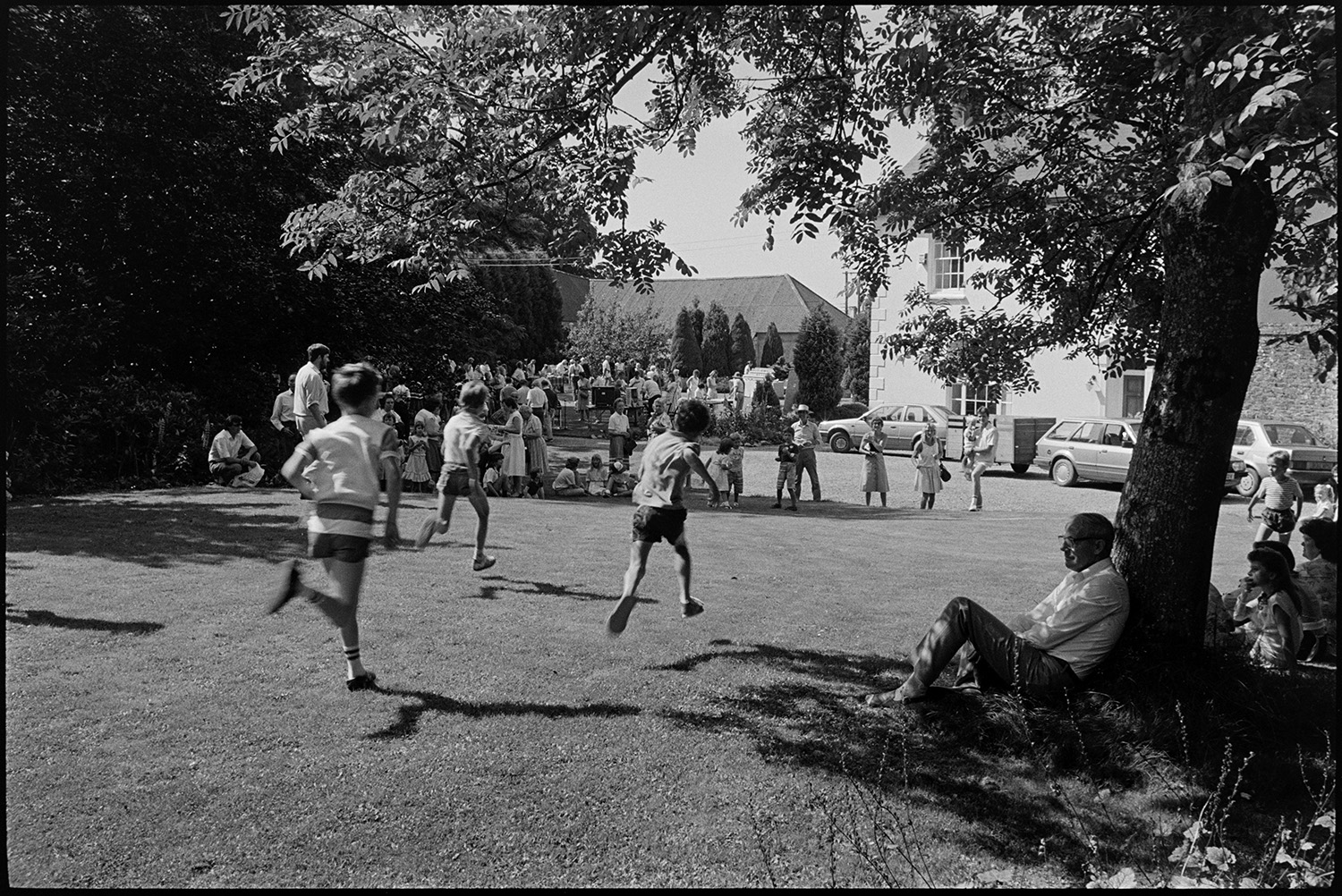 Vicarage garden fete, women spectators, games, dancing, man playing keyboard. Chatting!! 
[Children running in a race at Kings Nympton vicarage fete. A crowd of people are watching and a man is sat under a tree in the shade in the foreground.]