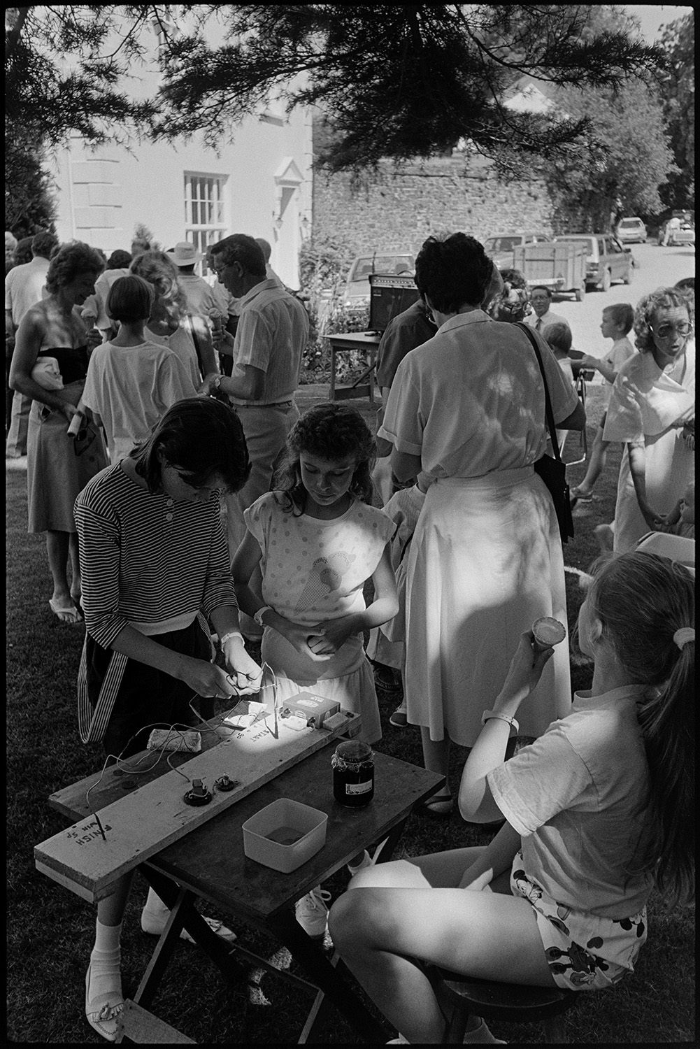 Vicarage garden fete, women spectators, games, dancing, man playing keyboard. Chatting!! 
[Children having a go on a buzzer game at Kings Nympton vicarage fete. A girl running the stall is eating an ice cream and other visitors to the fete can be seen in the background.]