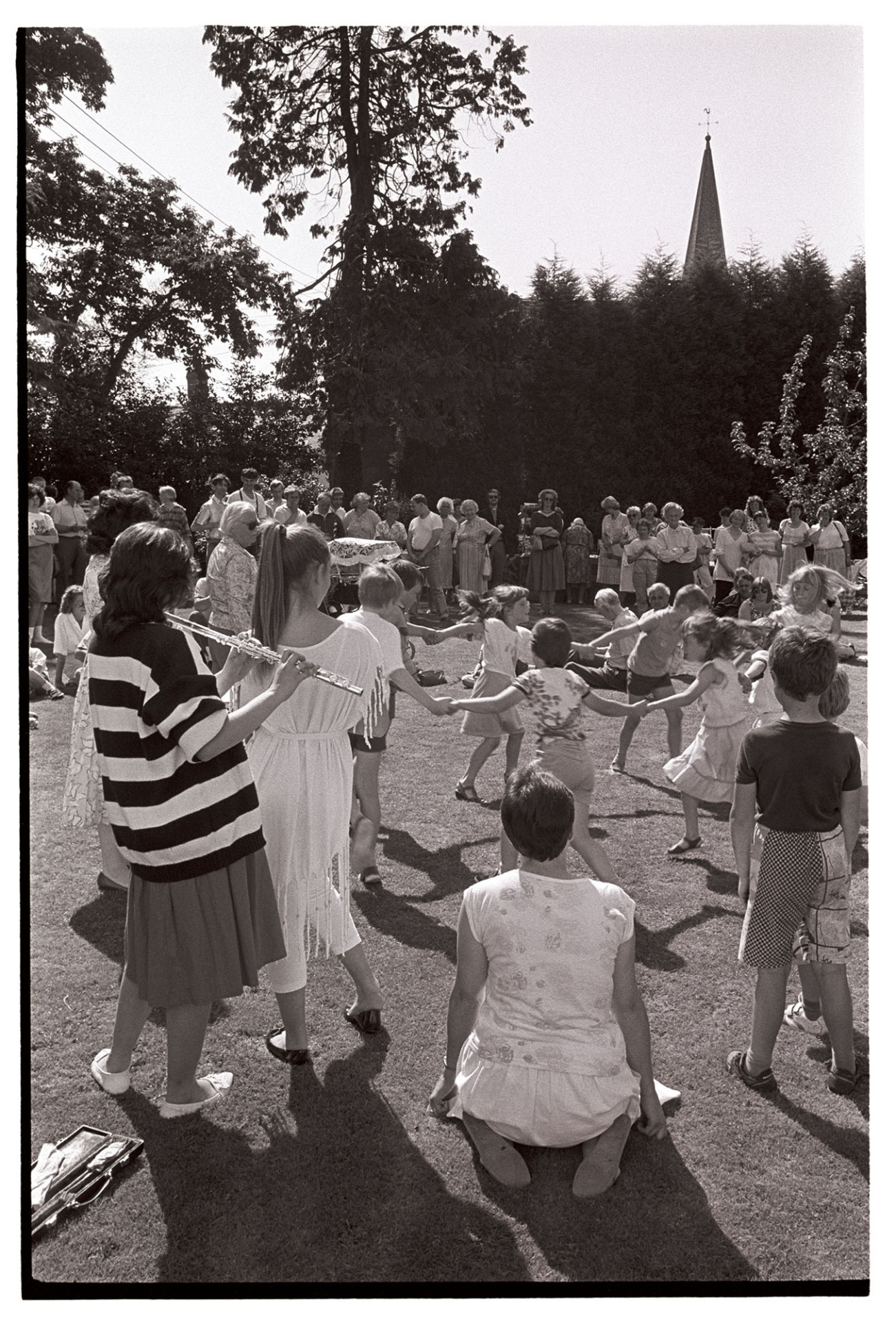 Vicarage fete, garden party children dancing to flute etc. 
[Children dancing in a circle to a woman playing a flute at the Vicarage fete at Kings Nympton. Spectators are watching in the background and the church spire can be seen behind the trees.]