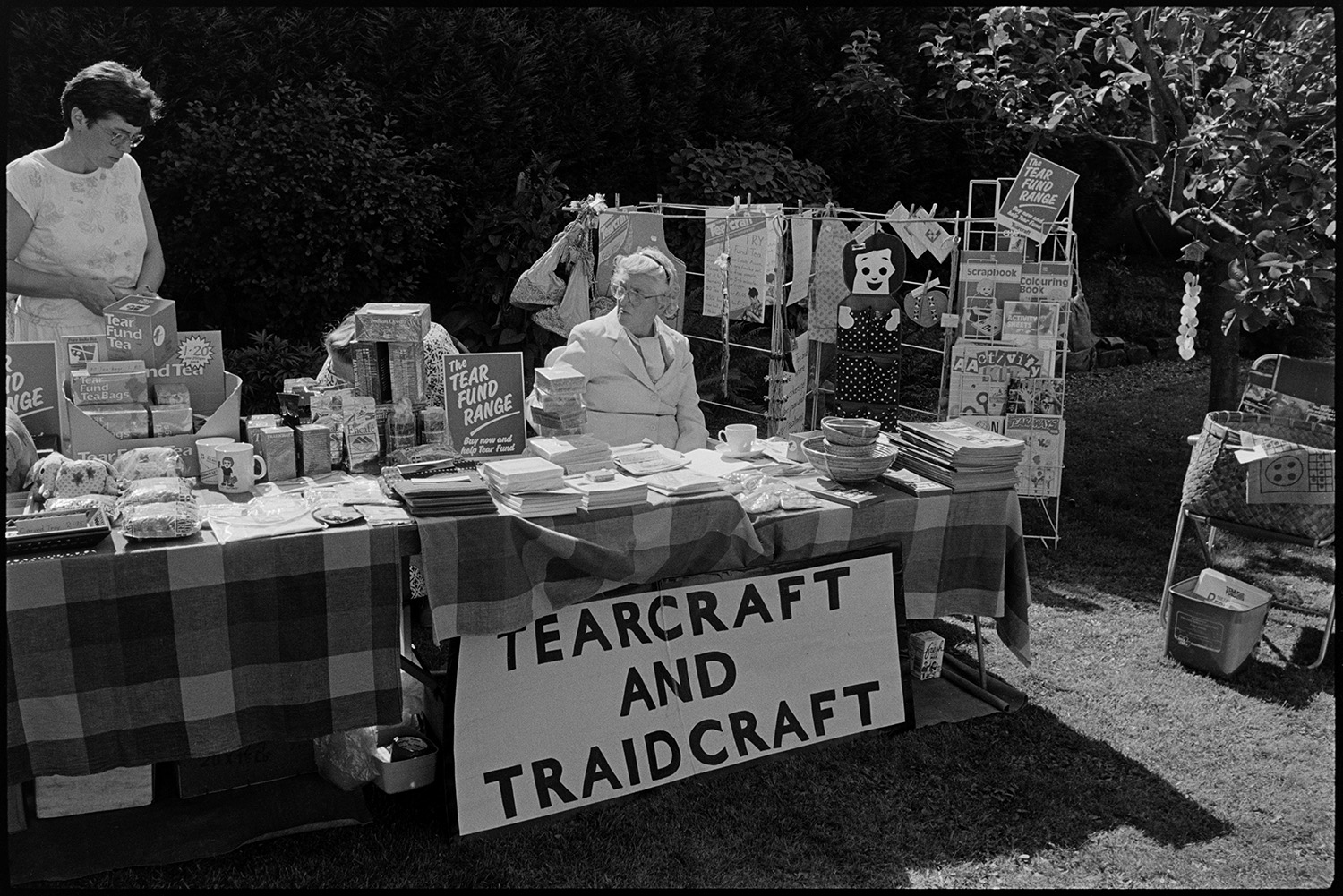 Vicarage fete, garden party people sitting at table with tea and cakes, women chatting. 
[Two women running a Tearcraft and Traidcraft stall at Kings Nympton vicarage fete. Various items are for sale including tea, mugs, books and baskets.]