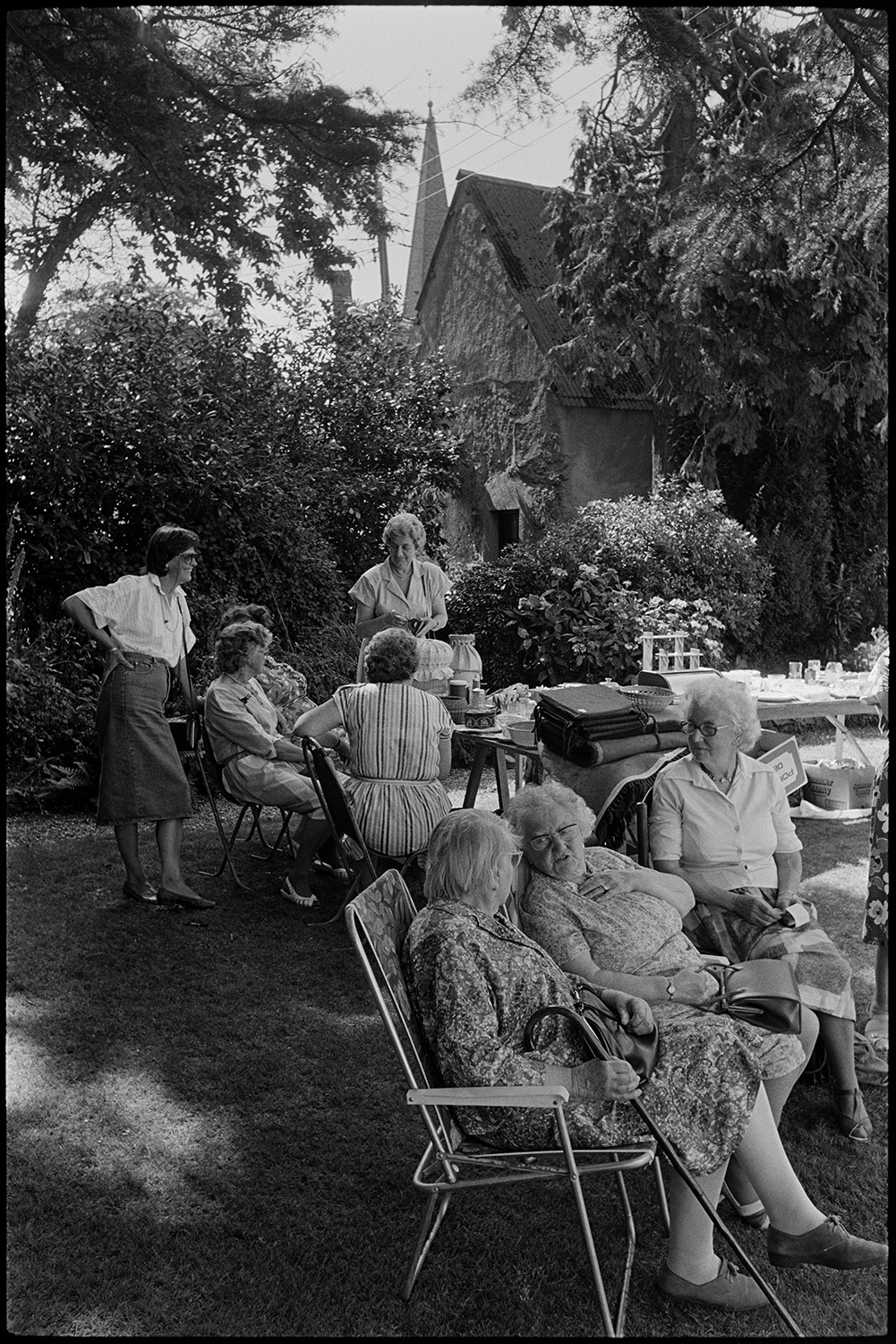 Vicarage fete, garden party people sitting at table with tea and cakes, women chatting. 
[Women sat on garden chairs and talking at Kings Nympton vicarage fete, in the shade of trees.]