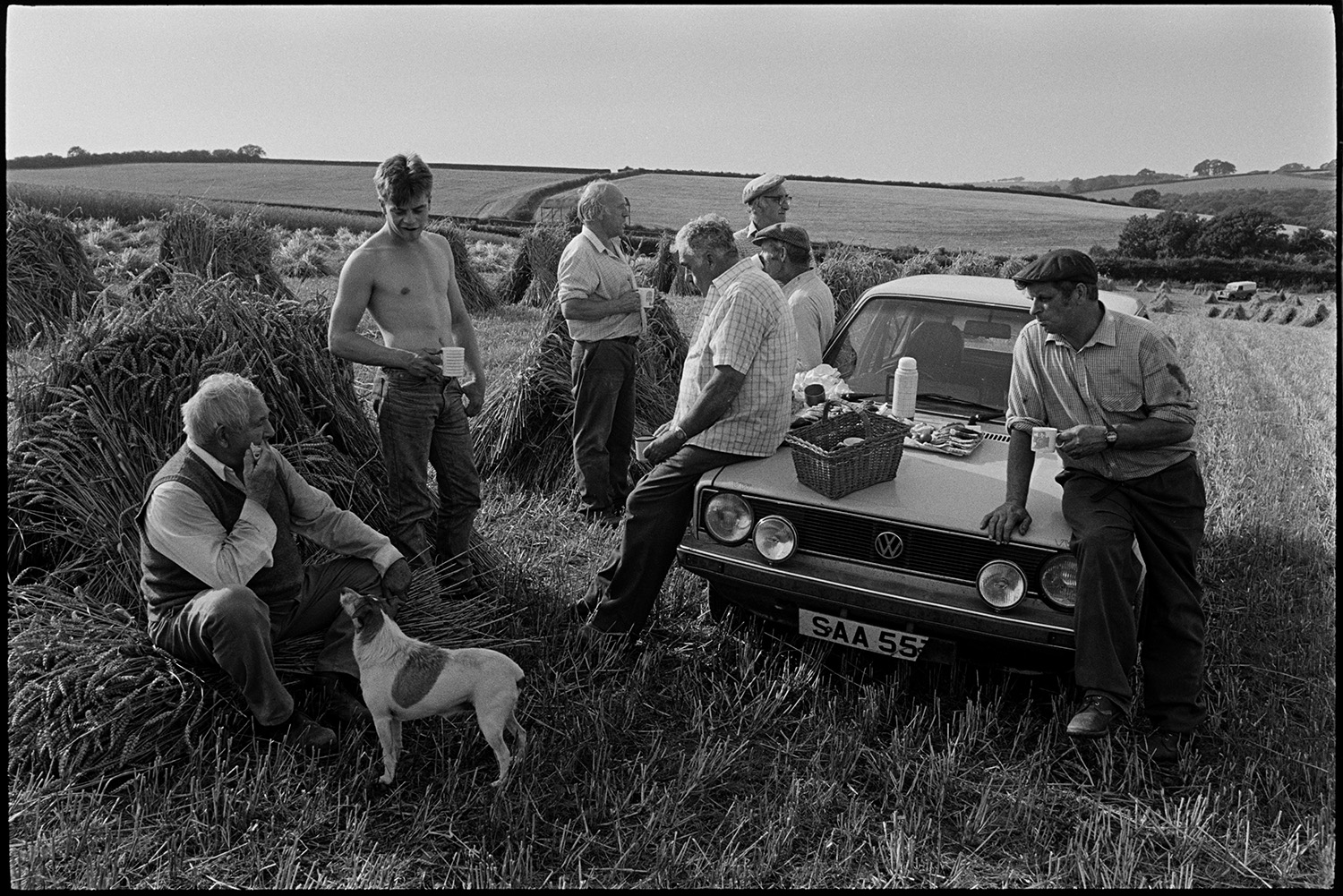 Harvesters tea break, leaning on car, woman, farmer's wife pouring from kettle, dog. 
[Seven men, possibly from the Down family, having a tea break from harvesting by a car, in a field at Spittle Farm, Chulmleigh. Cakes, a basket and thermos flask are on the bonnet of the car. A dog is with one man who is sat against a corn stook. Other corn stooks can be seen in the background.]