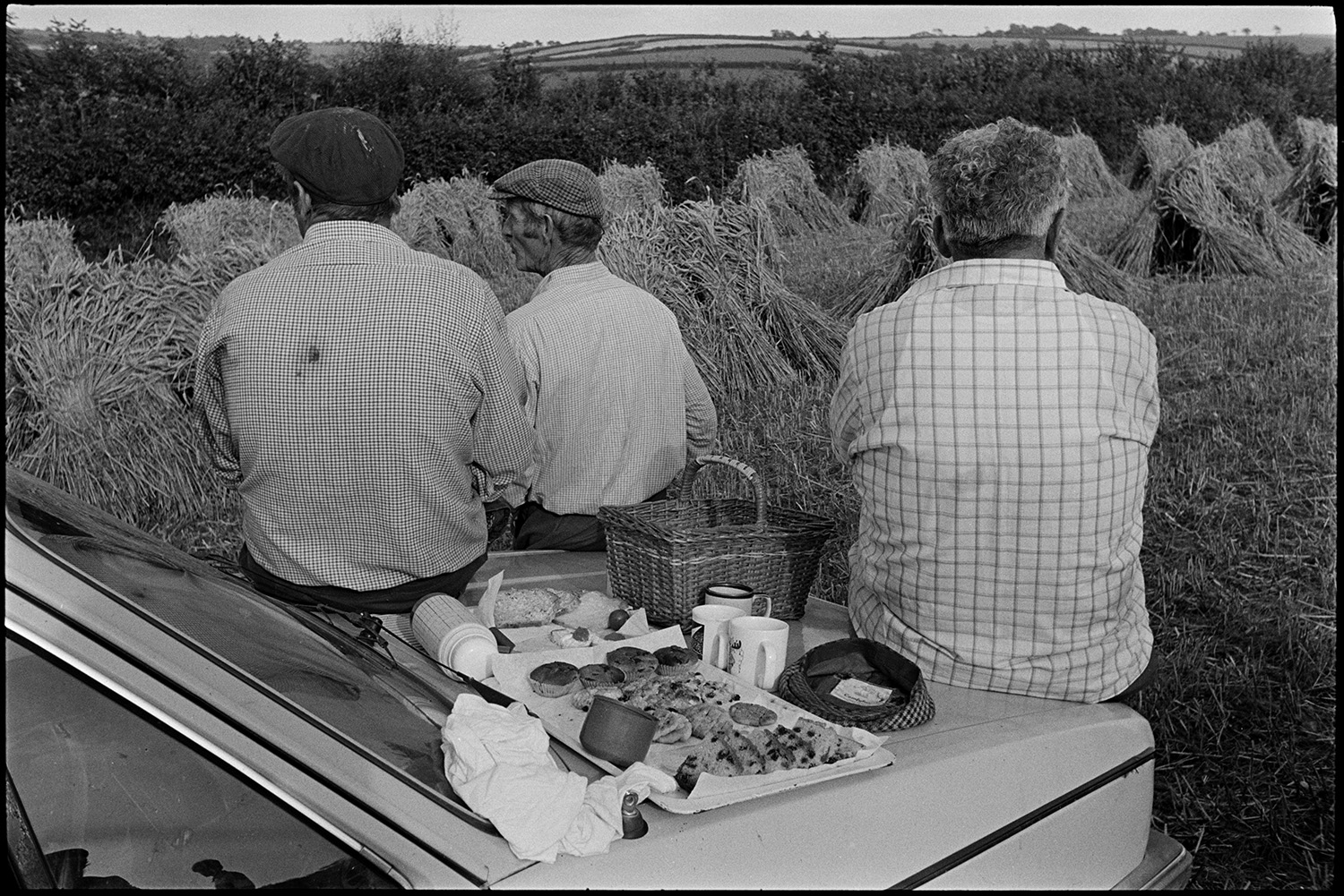Harvesters tea break, leaning on car, woman, farmer's wife pouring from kettle, dog. 
[Three men, possibly from the Down family, sat on the bonnet of a car having a tea break from harvesting in a field at Spittle Farm, Chulmleigh. Cakes, mugs, a thermos flask and a basket are also laid out on the car bonnet. Stooks of corn can be seen in the field in the background.]