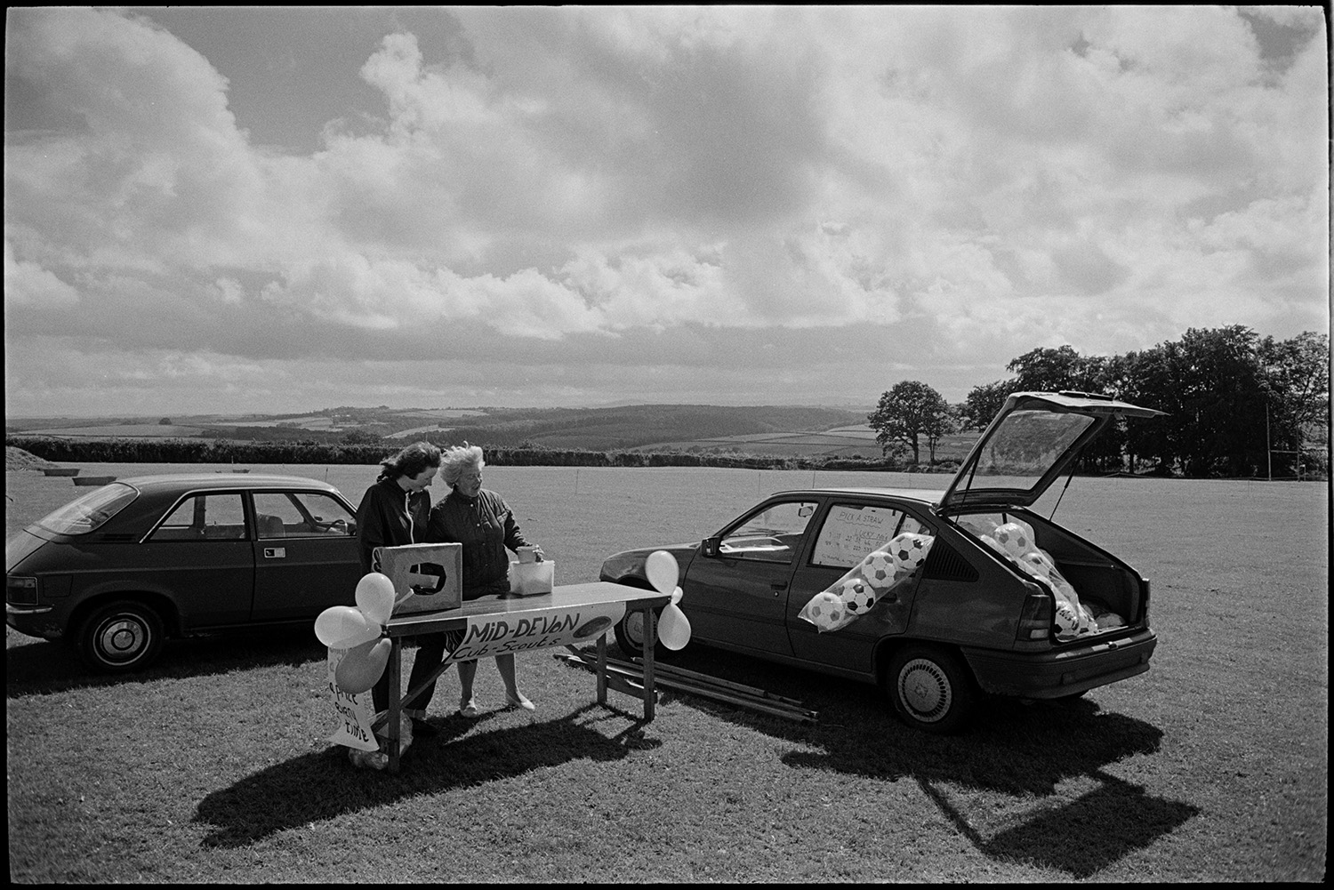 Sports Day, sheep dog trials, races, stalls, football. 
[Two women running a stall for the Mid-Devon Cub-Scouts at a sports day in Chulmleigh playing fields.  A car with nets of footballs is parked next to them.]