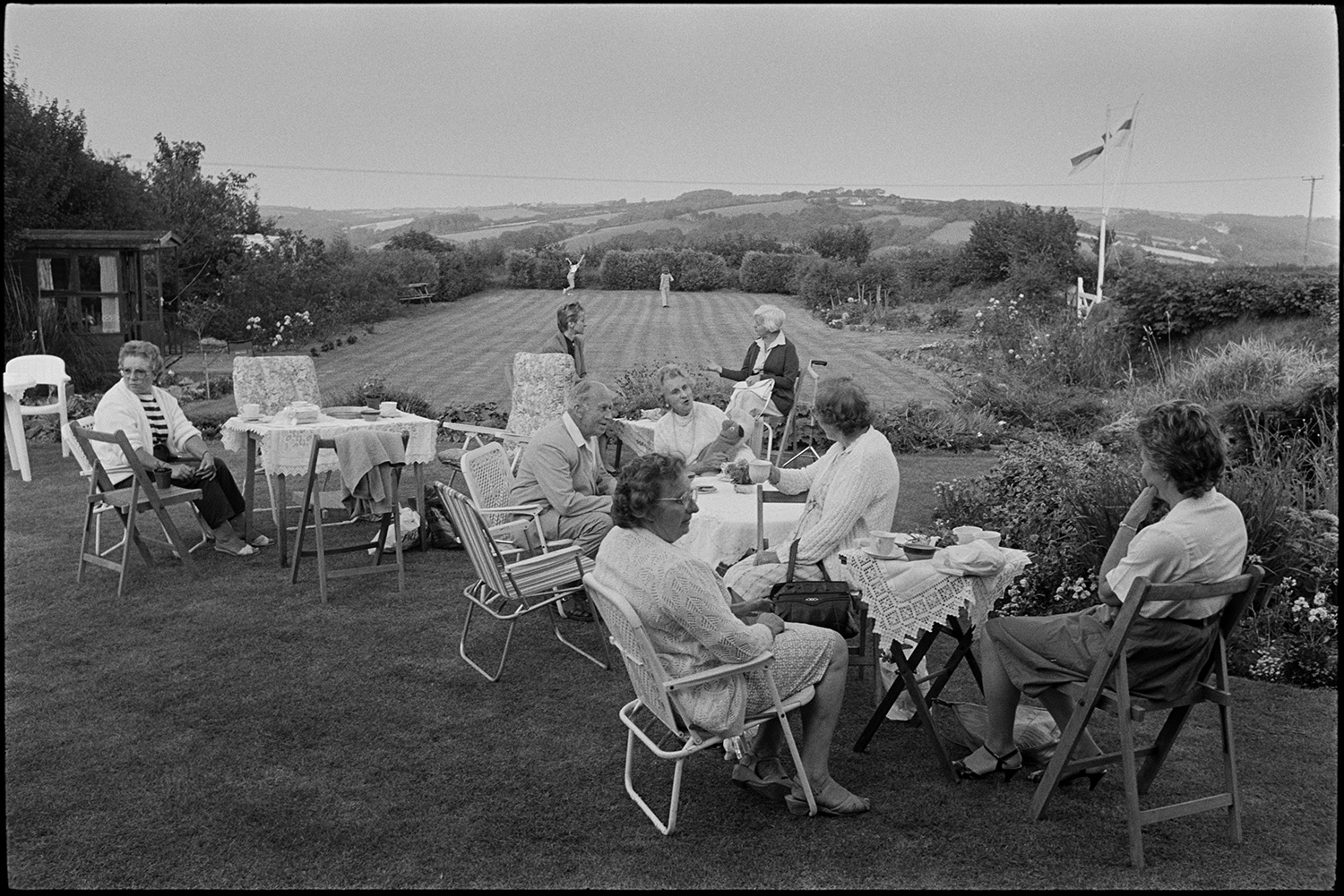 Garden fete, people having tea cake stall. 
[Men and women sat on garden chairs talking, drinking tea and eating cakes at a fete at Cricket Close, Chulmleigh. A Union Jack flag is flying from a flagpole and children are playing in the garden in the background.]