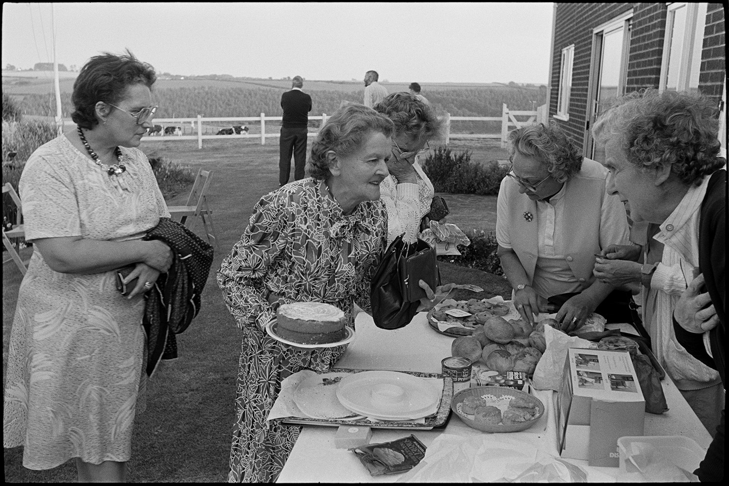 Garden fete, people having tea cake stall. 
[Women buying cakes from a cake stall at a fete at Cricket Close, Chulmleigh. Three men are watching a cricket match and cows grazing over a wooden fence in the background.]