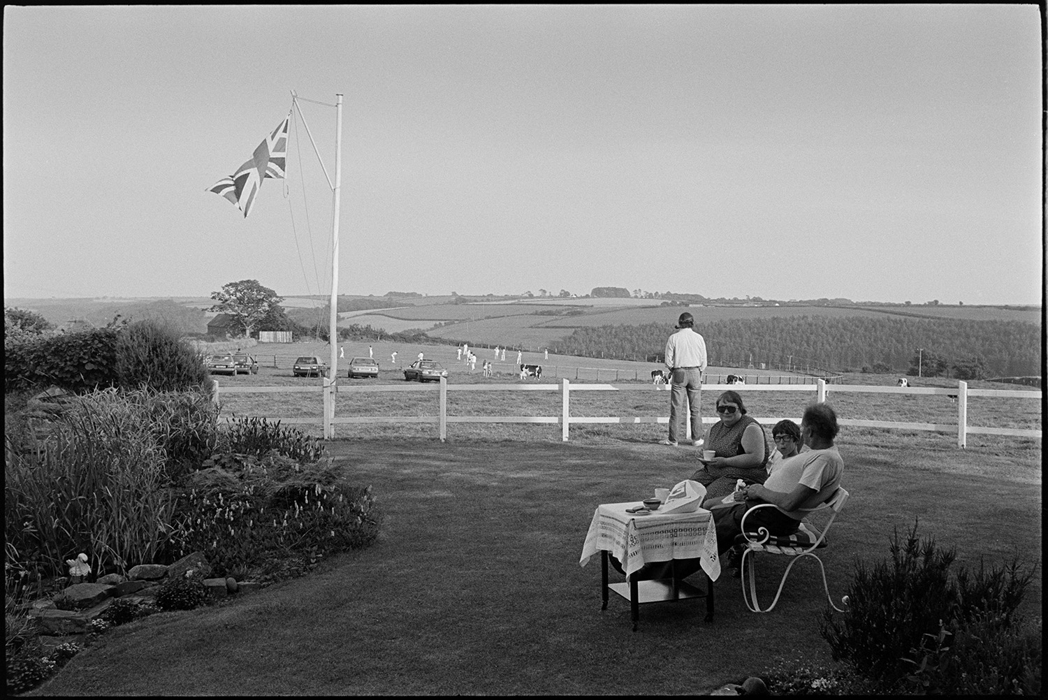 Garden fete, people having tea, cake stall. 
[Three people sat on a bench drinking tea and eating cakes at a fete at Cricket Close, Chulmleigh. A Union Jack flag is flying from a flagpole. In the background a man is stood looking over a wooden fence at a view of cows grazing and men playing cricket.]