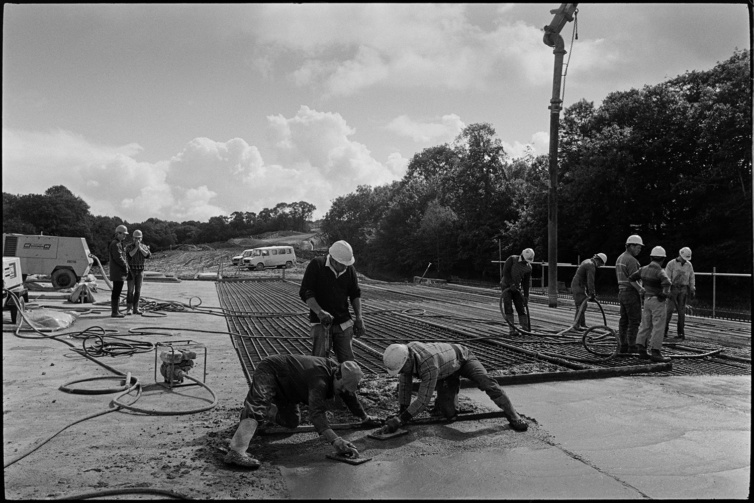 Men building new link road laying cement delivered by crane, laying polythene sheet. 
[Builders levelling newly laid cement over metal mesh on the Devon Link Road or A361 near South Molton. The cement is being delivered by a crane which is  visible by another group of builders on the right.]
