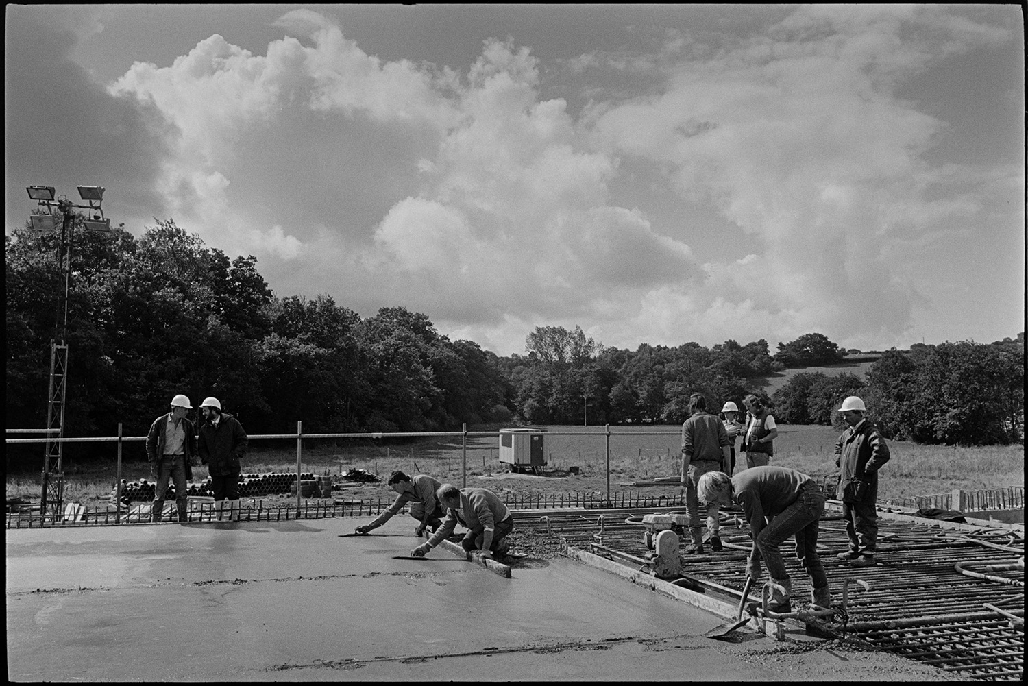Men building new link road laying cement delivered by crane, laying polythene sheet. 
[Builders levelling newly laid cement over metal mesh on the Devon Link Road or A361 near South Molton. They are using a shovel and a wooden baton. The cement was delivered by a crane.]