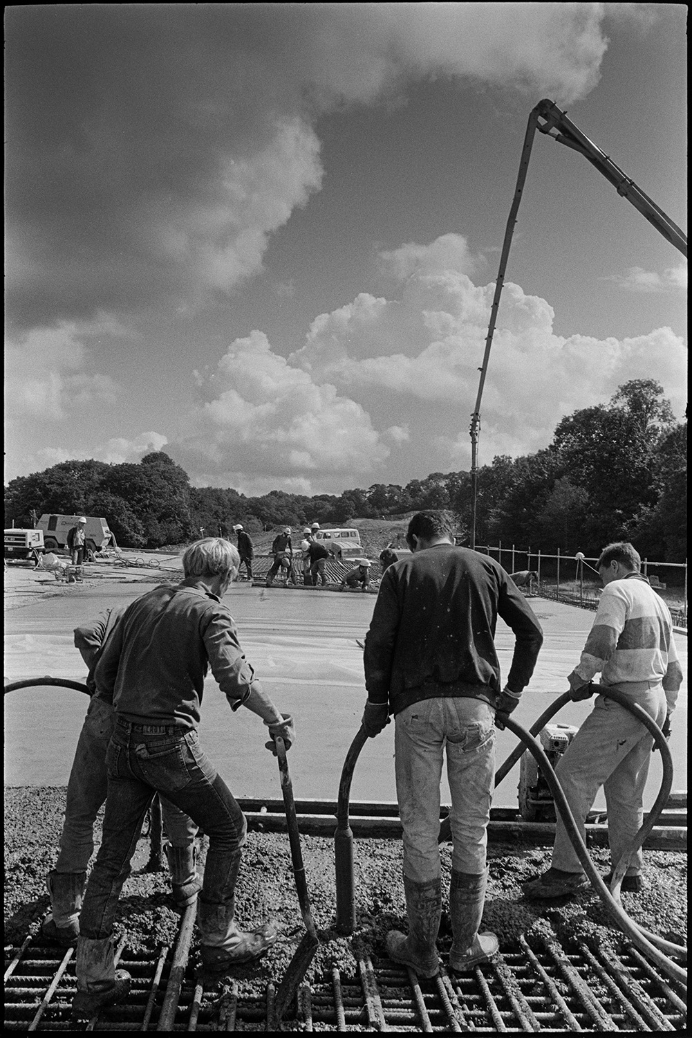 Men building new link road laying cement delivered by crane, sun and cloud. 
[Four men laying cement over metal mesh on the Devon Link Road or A361. The cement is being delivered by a crane. Other workmen can be seen in the background.]