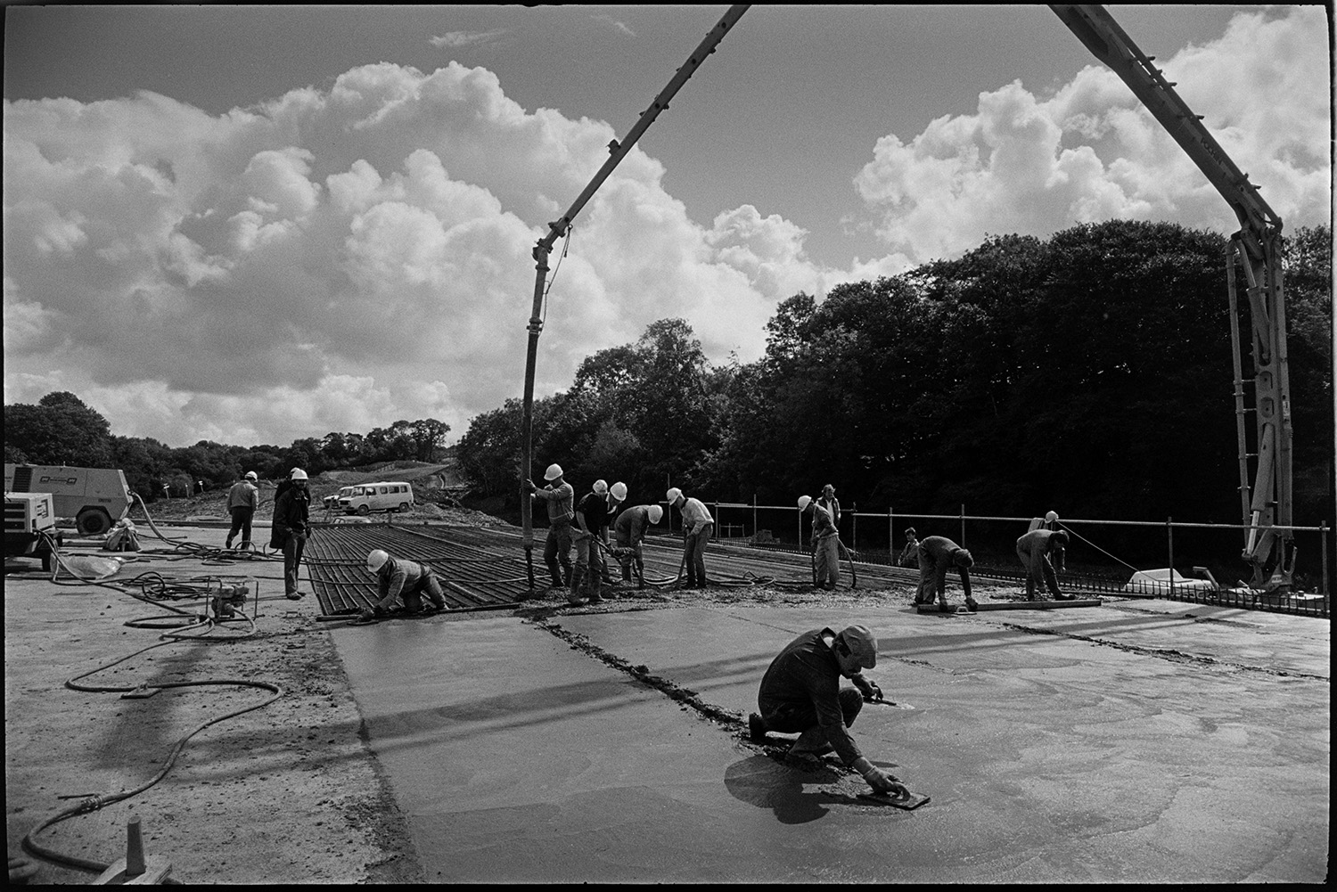 Men building new link road laying cement delivered by crane, sun and cloud. 
[Men laying cement over metal mesh on the Devon Link Road or A361. The cement is being delivered by a crane. A man is the foreground is levelling the cement.]
