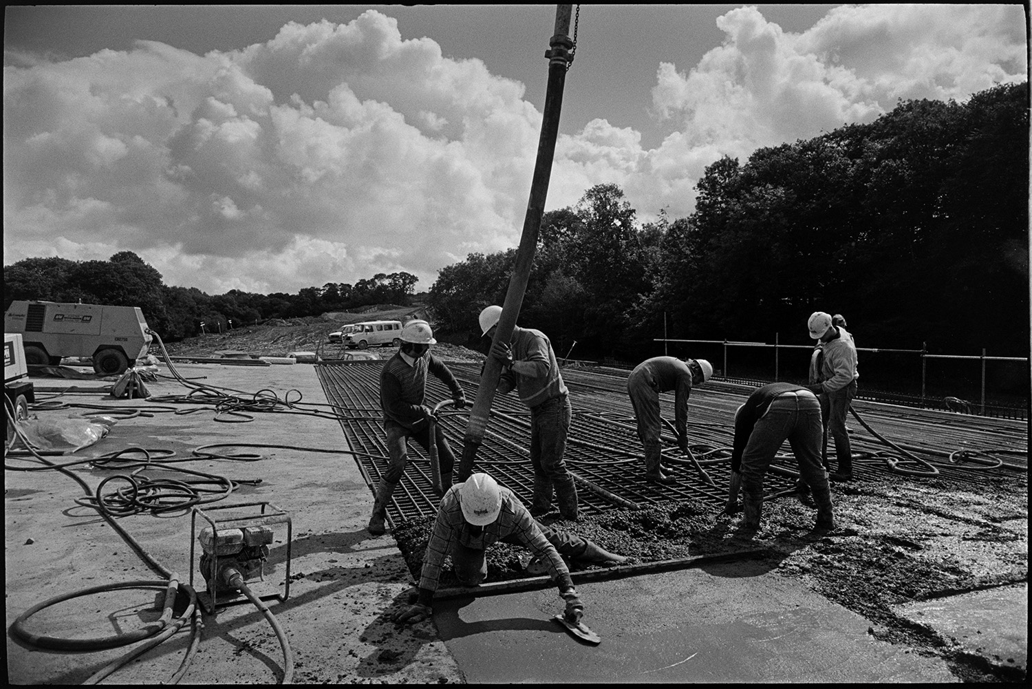 Men building new link road laying cement delivered by crane, sun and cloud. 
[Men laying cement over metal mesh on the Devon Link Road or A361. The cement is being delivered by a crane. A man is the foreground is levelling the cement with a wooden baton.]