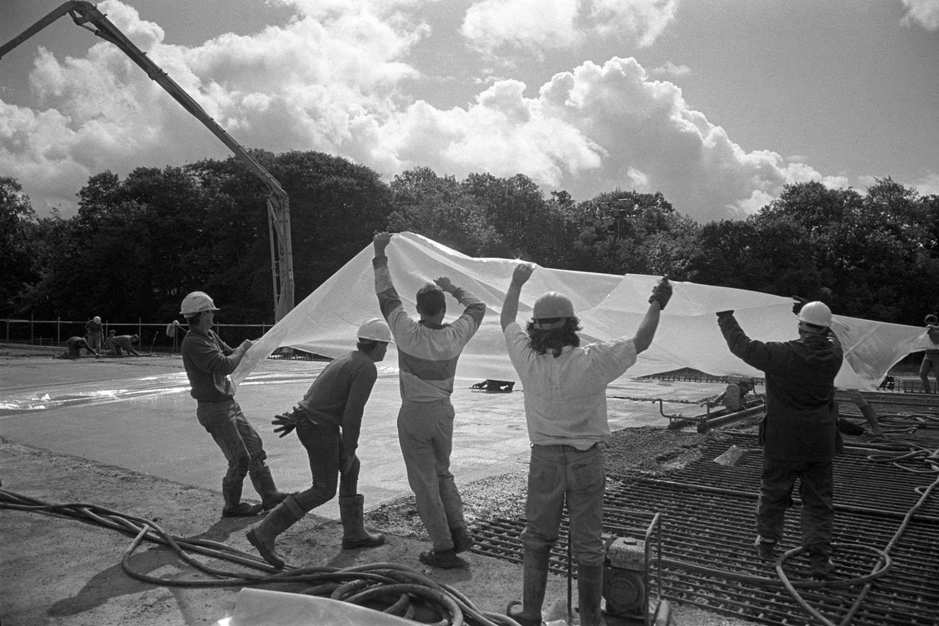 Men building new link road laying polythene sheet on top of wet cement, bright sun. 
[Men laying a polythene sheet over newly laid concrete on the new North Devon Link Road or A361 near South Molton. The crane which pumped in the concrete is visible in the background.]