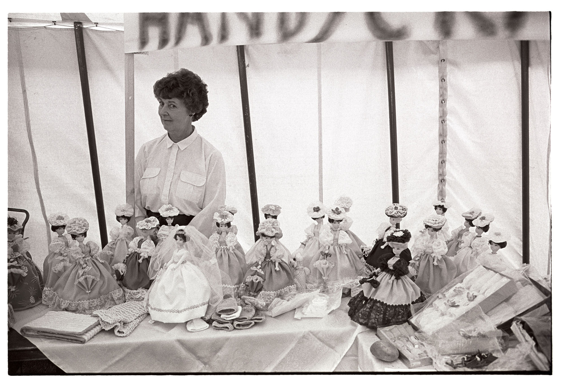 Fete, stall with dolls for sale, woman standing in tent. 
[A woman standing under a tent or marquee, running a stall selling dolls at a fete at Little Silver, High Bickington.]