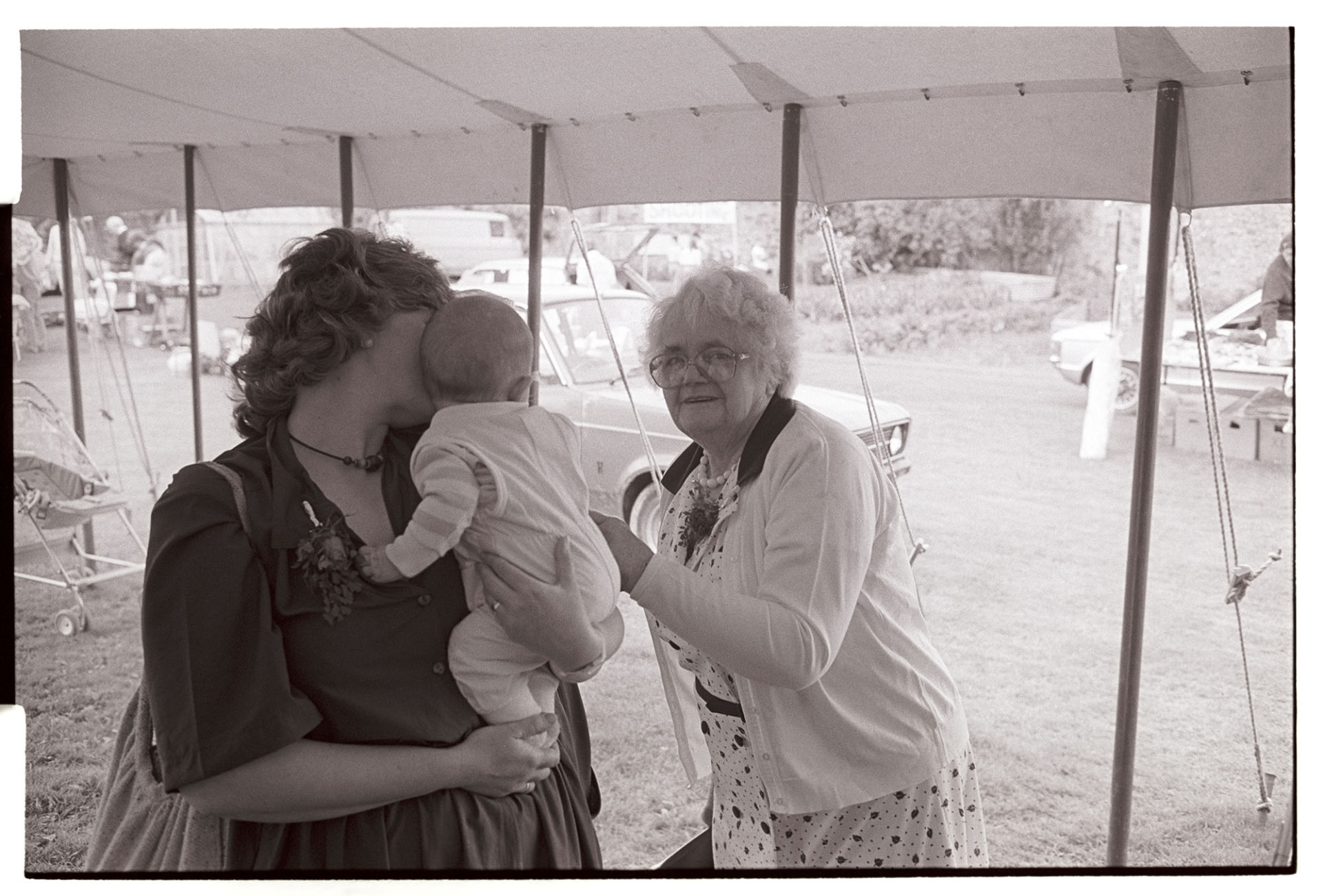 Elderly woman admiring baby. 
[A woman talking to a baby being held by another woman under a tent or marquee, at a fete at Little Silver, High Bickington.]