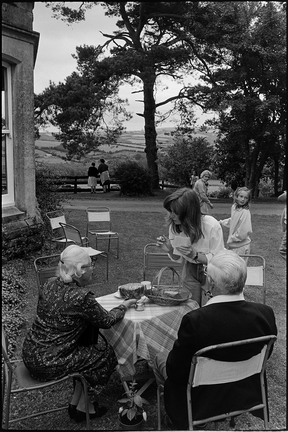 Fete with stalls, people having tea, looking at view, hand bell ringers. 
[A woman talking to a man and woman sat at a table at a fete at Little Silver, High Bickington. The woman seated is holding a cake. Other women and children can be seen on the lawn in the background.]