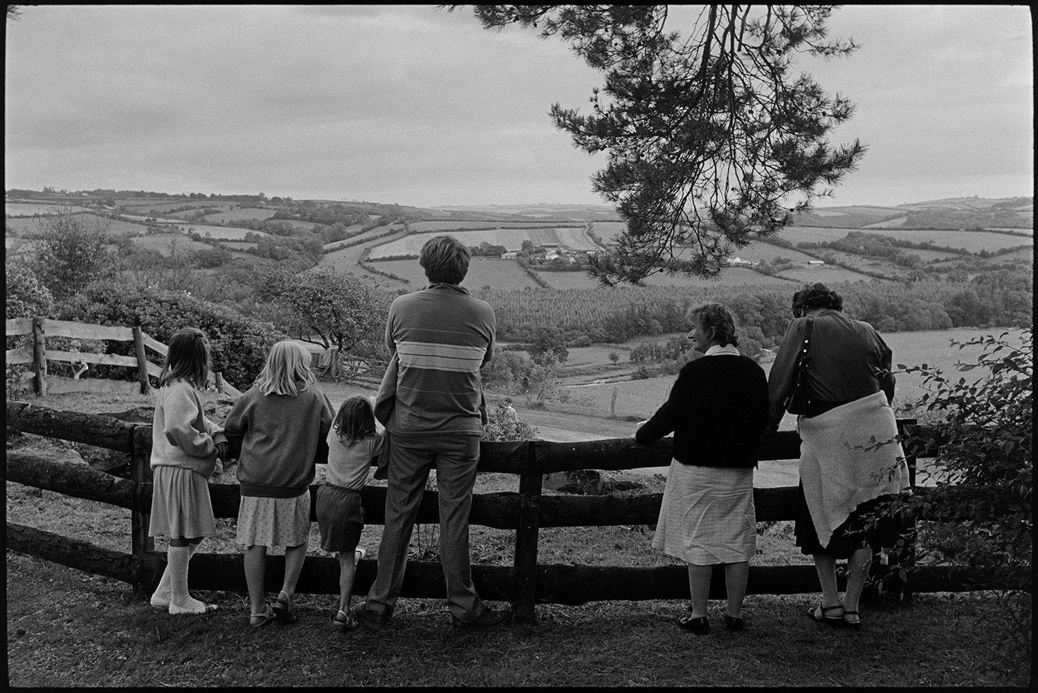 Fete with stalls, people having tea, looking at view, hand bell ringers. 
[Two women, a man and three children looking over a wooden fence to a view of fields, hedgerows and trees, at a fete at Little Silver, High Bickington.]