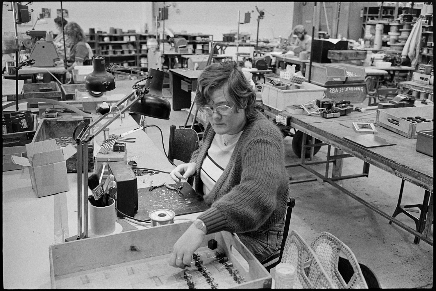 Inside transformer factory men and women making, designing transformers, computer. 
[A woman making transformers at Apex Transformers at Pathfields Business Park in South Molton. Other people can be seen working at desks in the factory in the background.