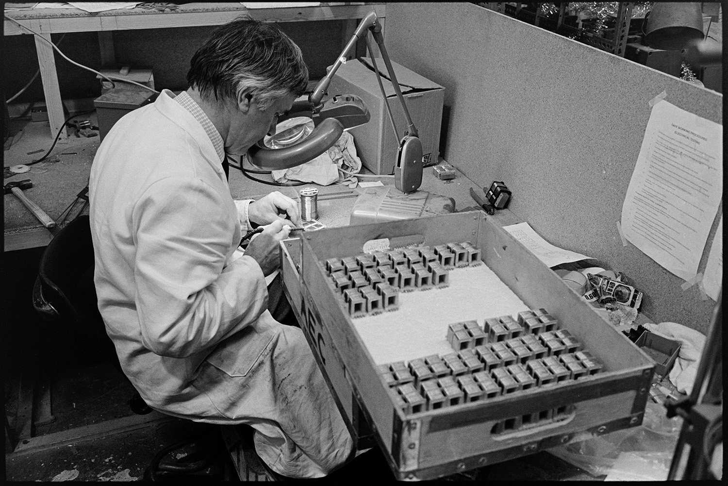 Inside transformer factory men testing, making, designing transformers, computer. 
[A man working with transformers at Apex Transformers at Pathfields Business Park in South Molton. He is using a magnifying glass.]