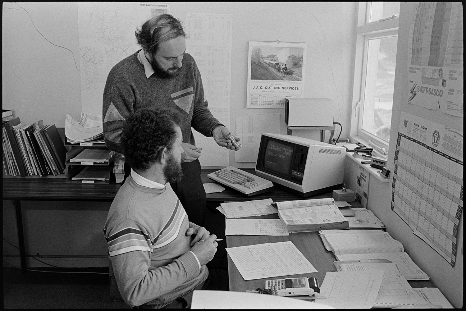 Directors of transformer factory in office working and using computer. 
[Two men talking and looking at a reel of wire at a desk in the office at Apex Transformers at Pathfields Business Park, South Molton. A computer is on the desk beside them.]