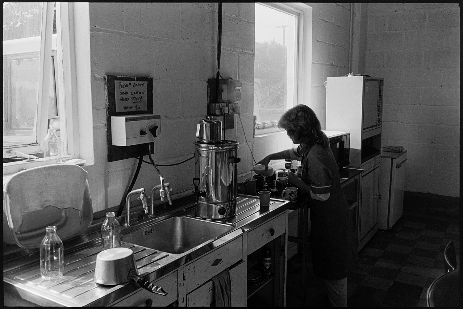 Inside transformer factory men testing, making, designing transformers, canteen sink. 
[A woman adding milk to cups of tea or coffee in the canteen at Apex Transformers at Pathfields Business Park, South Molton. A sink, urn, microwave and fridge can be seen in the canteen.]