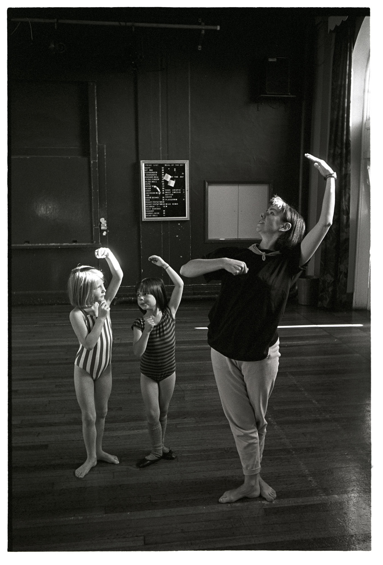 Children's dance class, teacher demonstrating to two small girls. 
[Sally Barber teaching two young girls a dance or ballet class a Chulmleigh Community College.]