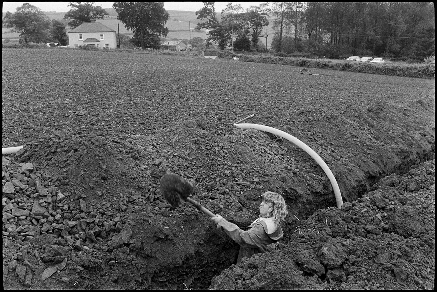 Farmer and daughter, woman, laying drainage pipe in ploughed field, tractor, JCB. 
[Rob Millman's daughter digging in a trench to lay a drainage pipe in a ploughed field near Leigh Cross, Chulmleigh. A house and parked cars are visible in the background.]