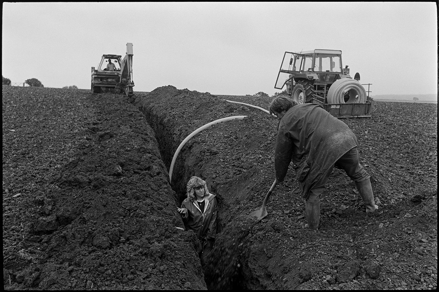 Farmer and daughter, woman, laying drainage pipe in ploughed field, tractor, JCB. 
[Rob Millman and his daughter laying a drainage pipe in a ploughed field near Leigh Cross, Chulmleigh. His daughter is stood in the trench with the pipe while Rob Millman shovels earth in to cover the pipe. A JCB can be seen in the background digging a trench for more pipework. A tractor with a link box containing another roll of drainage pipe in also visible.]