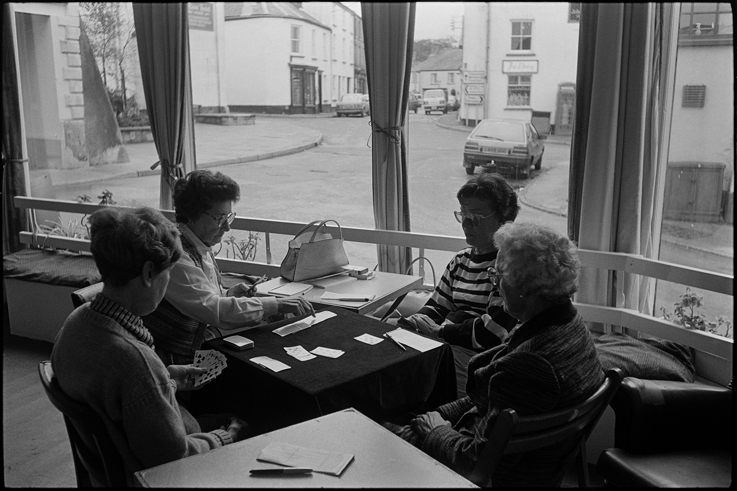 Women playing cards in social club, 61 centre. 
[Four women playing cards at a table in the 61 Centre in Chulmleigh. A street can be seen outside with parked cars and shop fronts.]