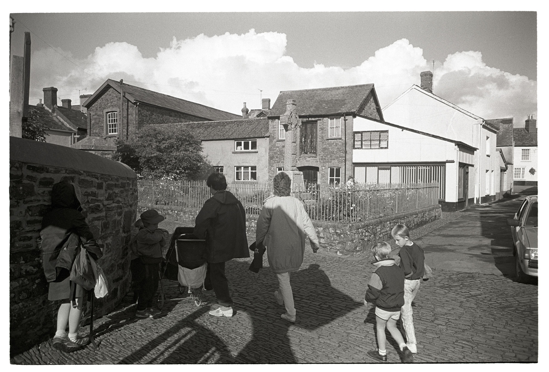 Women, mothers and children after school. clouds in background. 
[Mothers and children walking home from school along a cobbled street in Chulmleigh. They are walking past the war memorial. Clouds can be seen in the sky in the background.]