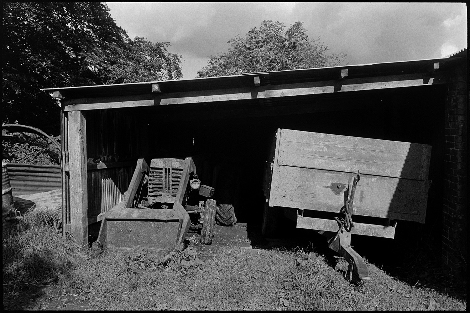 Dog barking in farm lane with parked harrow, old bulldozer. 
[An old tractor and wooden trailer parked in a shed at Spittle Farm, Chulmleigh.]