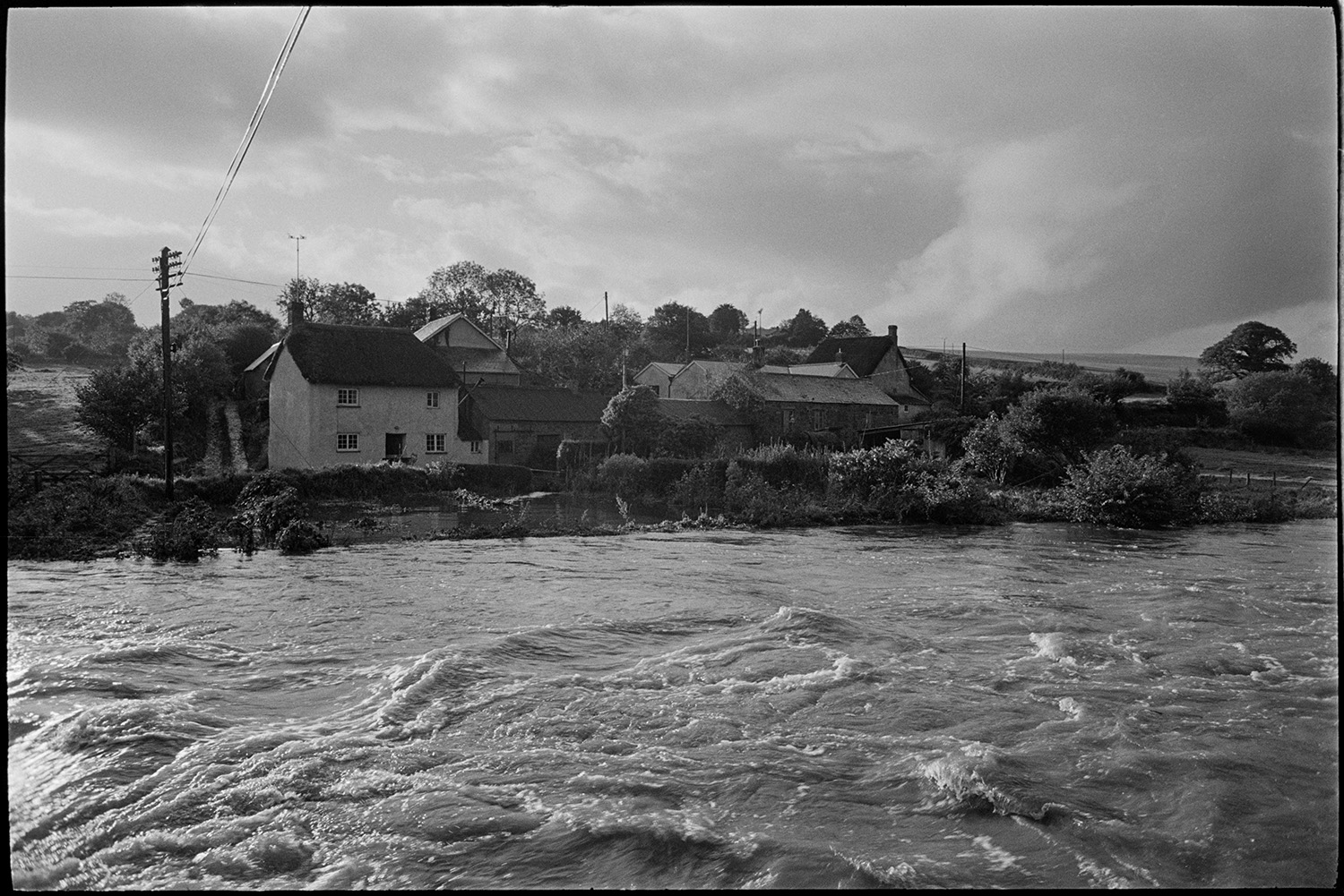 River in flood, marooned thatched cottage, iron gate in flooded field. 
[Cottages cut of by flooding from the River Taw at Bridge Reeve, Ashreigney.]