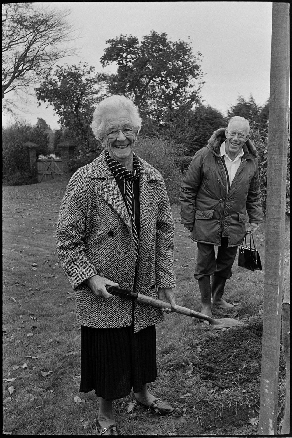 Tree planting at Arts Centre. 
[Mrs Branhan and Gareth Keen planting at tree at the Beaford Centre at Greenwarren House in Beaford. Gareth is holding the woman's handbag while she shovels earth around the tree.]