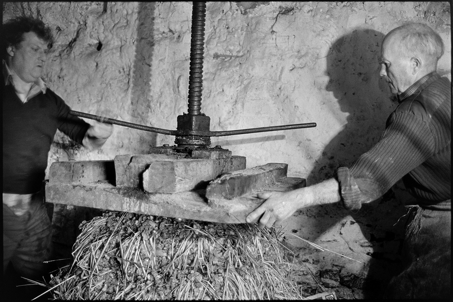 Orchards, Devon, man operating cider press, making cheese. 
[Bill Hammond, on the left, and another man operating a cider press in a barn at Rashleigh Mill, Bridge Reeve. It is compressing the cheese which is layers of apple pulp and straw.]