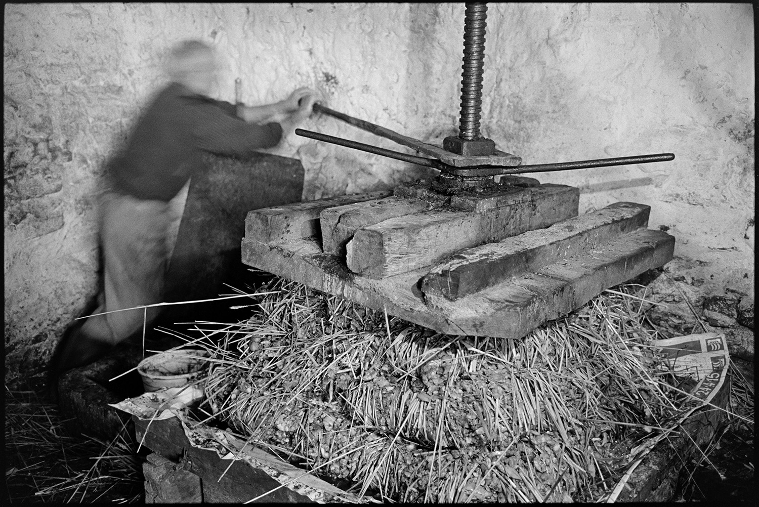 Orchards, Devon, man operating cider press, making cheese. 
[Bill Hammond operating a cider press in a barn at Rashleigh Mill, Bridge Reeve. He is compressing the cheese, which is layers of apple pulp and straw.]