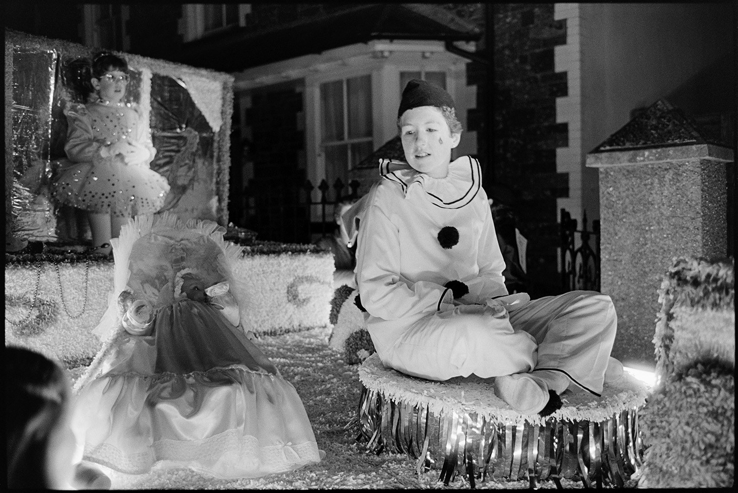 Carnival floats at night majorettes looking on. 
[Children in fancy dress on a carnival float at Dolton carnival at night. One of them is dressed as a pierrot character.]