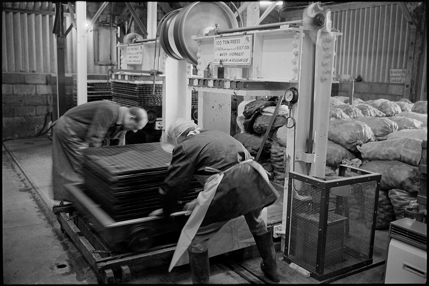 Cider making, men operating cider press in factory. 
[Two men working with a 100 ton cider press built in 1936, at Hancock's cider factory at Clapworthy Mill, South Molton. Sacks of apples can be seen stacked in the background.]