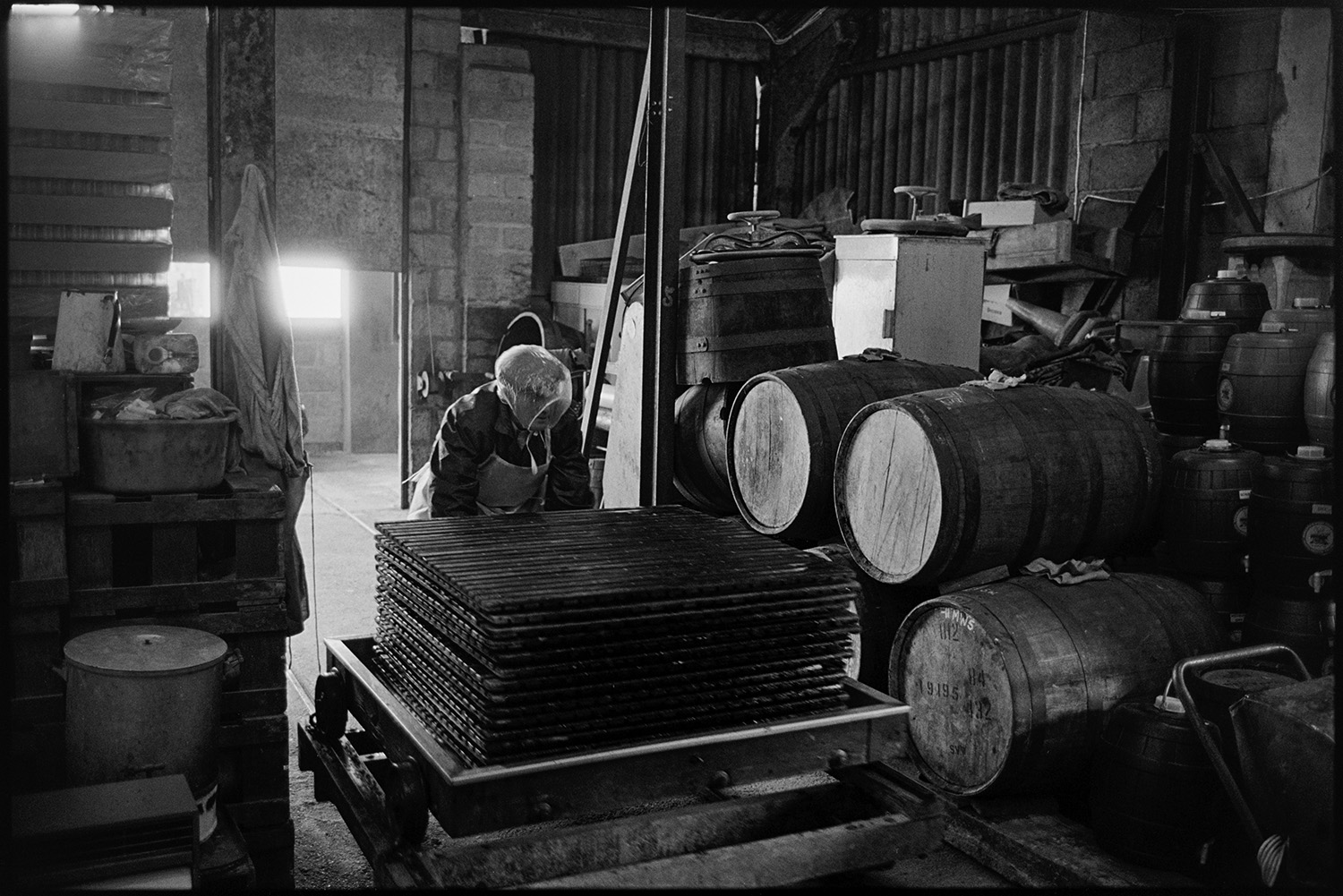 Cider making, men operating cider press in factory. 
[A man moving a cider press past barrels of cider at Hancock's cider factory at Clapworthy Mill, South Molton. He is wearing a polythene hat.]