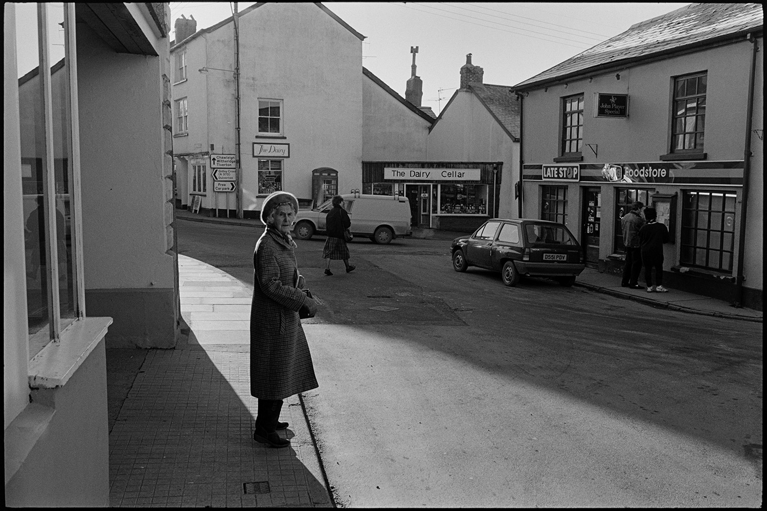 Street scene, early morning. 
[People shopping in Fore Street, Chulmleigh in the early morning. A woman is stood on the pavement in the foreground and two people are looking at a noticeboard outside the shopfront of Late Stop Food store. The shop front of The Dairy Cellar is also visible in the background.]